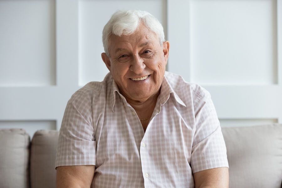 Smiling man seated indoors on a sofa, wearing a light plaid shirt.