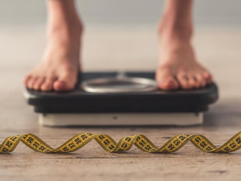 Feet on a bathroom scale beside a yellow measuring tape on a wooden floor.