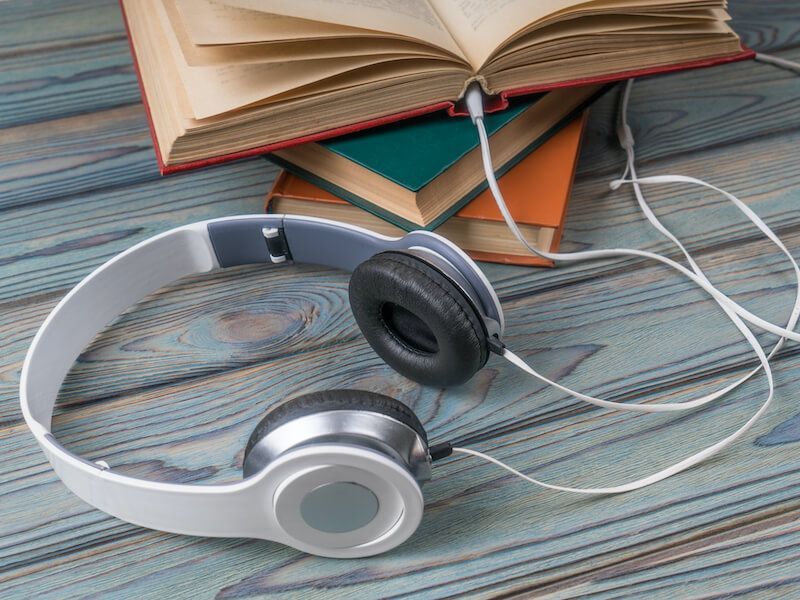 Silver and black headphones beside an open book on a wooden desk, with tangled white earbud cords