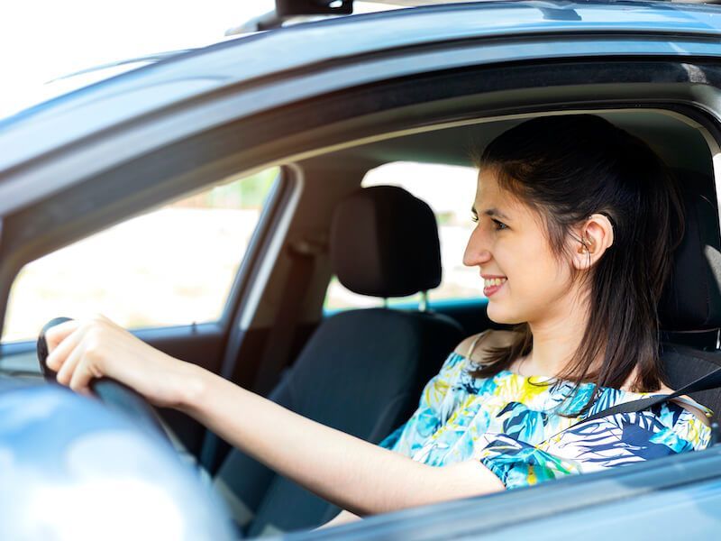 Woman smiling while driving a car, wearing a blue patterned shirt.