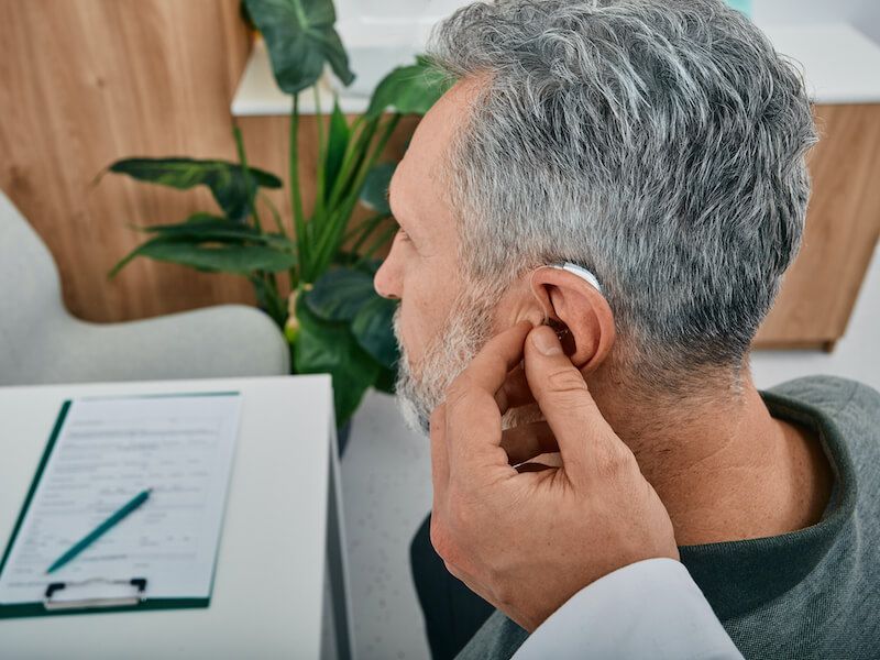 Man adjusting a hearing aid at a desk with paperwork and a plant in the background