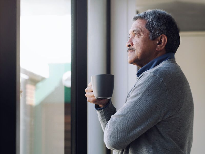Man in a gray sweater holding a mug and looking out a window in a bright room