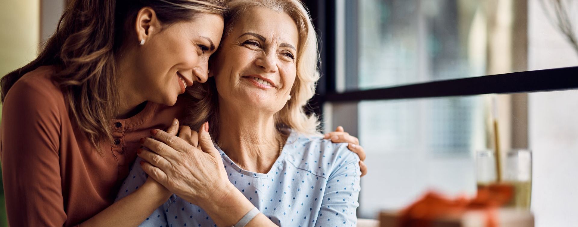 Two women sit close together indoors, smiling and embracing near a window.