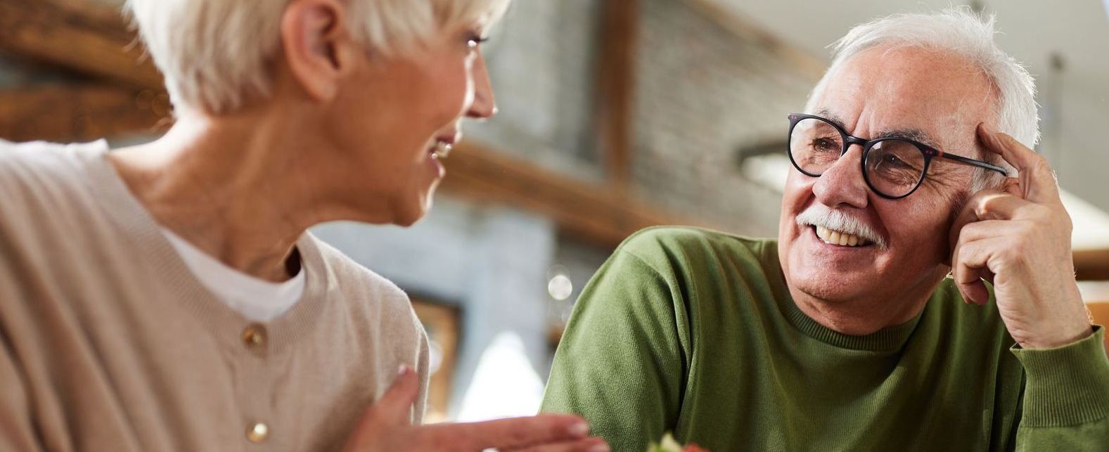 Two older adults smiling and talking indoors, one in a green sweater and glasses
