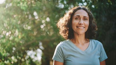 Smiling person in a light blue T-shirt standing outdoors with greenery in the background