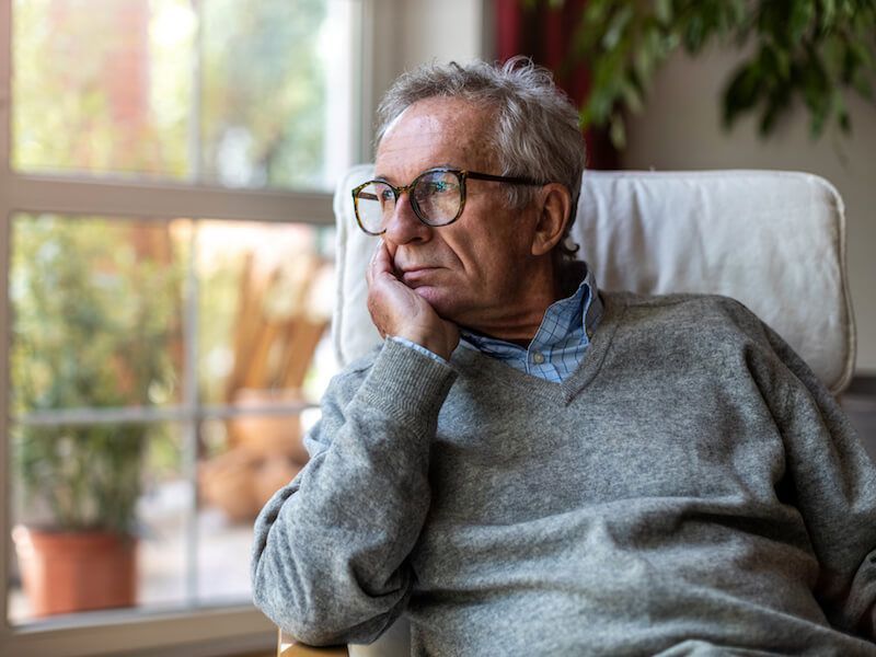 Older man in glasses and gray sweater sits by a window, looking thoughtfully outside.