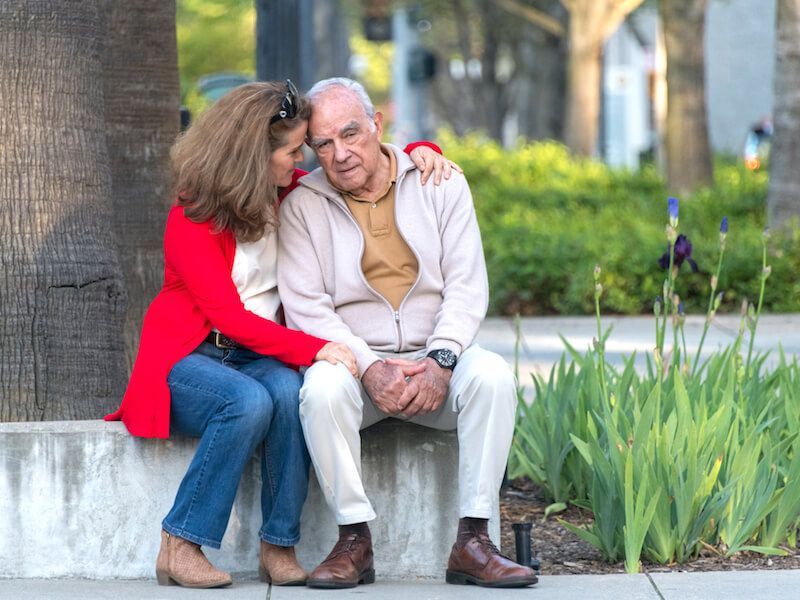 Couple sitting on a bench in a park, woman in red sweater leaning toward man in beige sweater.