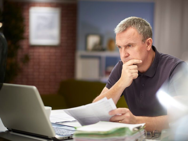 Man studying papers beside a laptop at a desk, looking thoughtful in an office.