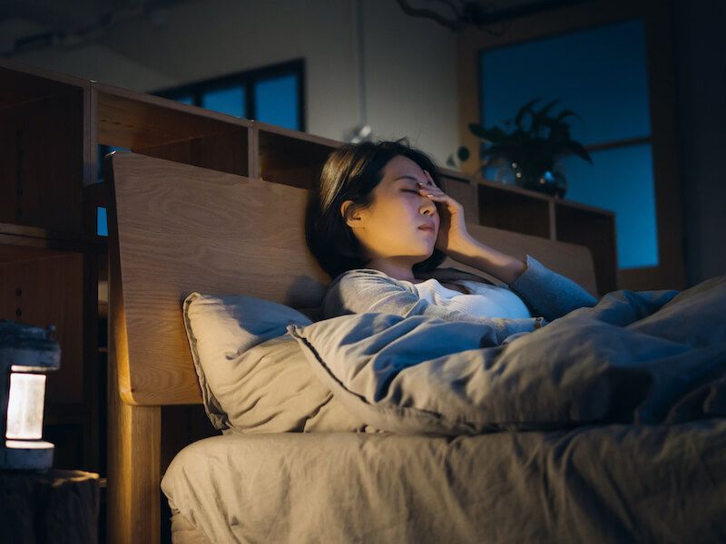 Person lying in bed with a hand on their face, lit by cool blue light at night