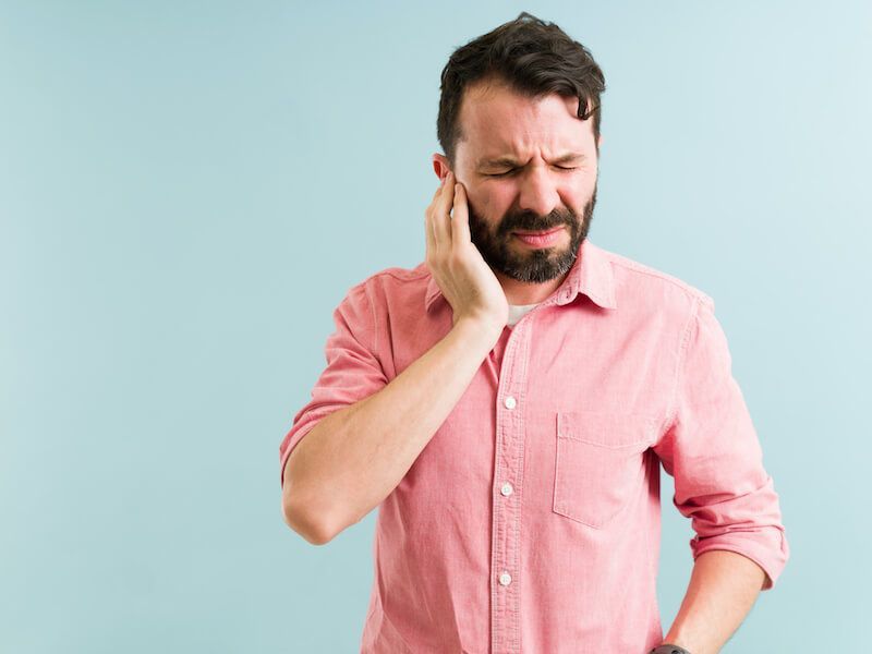 Man in pink shirt holding his cheek and wincing against a light blue background