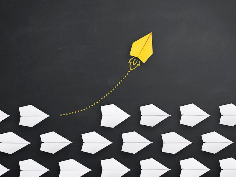Yellow paper airplane flying above a scattered flock of white paper planes on a dark background