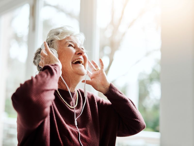 Smiling elderly woman wearing headphones and a maroon sweater, enjoying music by a bright window