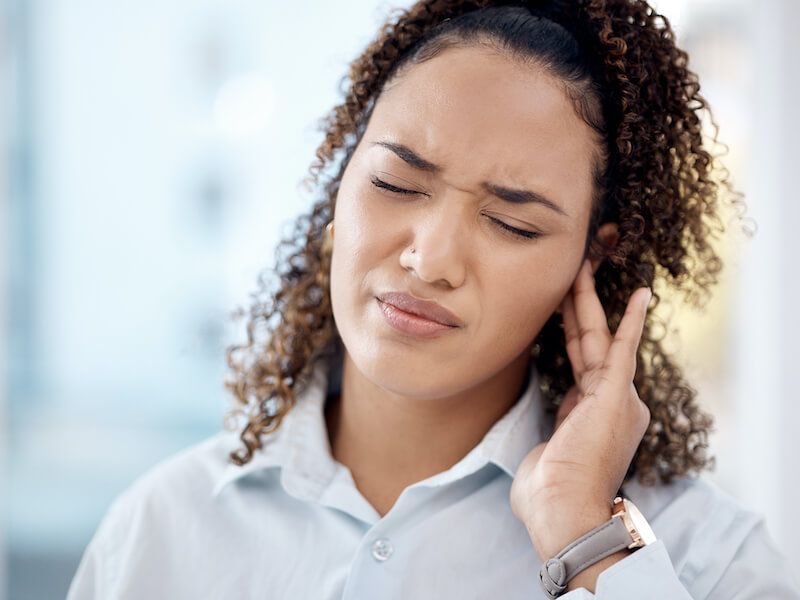 Woman with eyes closed, holding her ear in discomfort, wearing a white shirt and smartwatch.