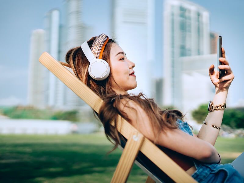 Woman wearing white headphones relaxing in a wooden chair and looking at her phone in a sunny park