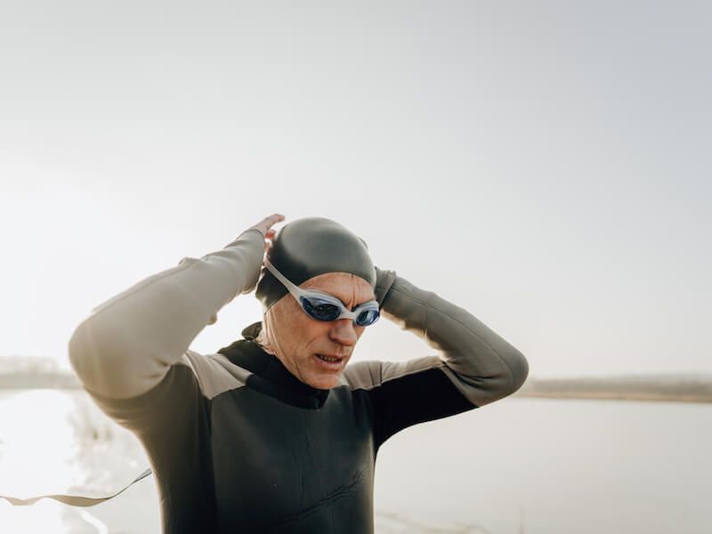 Swimmer in black wetsuit and blue goggles adjusting cap at the water’s edge