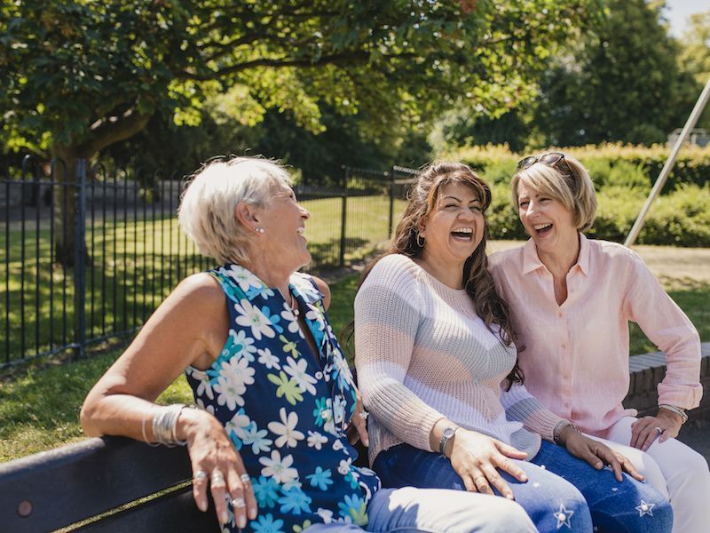 Three women laughing on a park bench in sunny greenery
