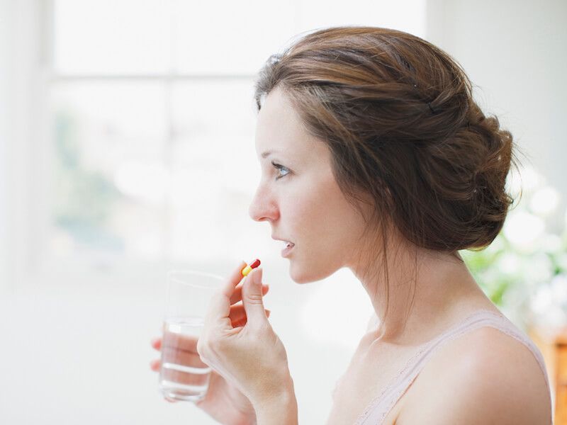Woman in profile holding a glass of water and a pill near her mouth in a bright room