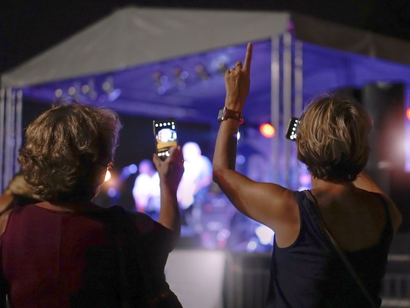 Two people at an outdoor concert, one filming with a phone and the other raising an arm under purple stage lights