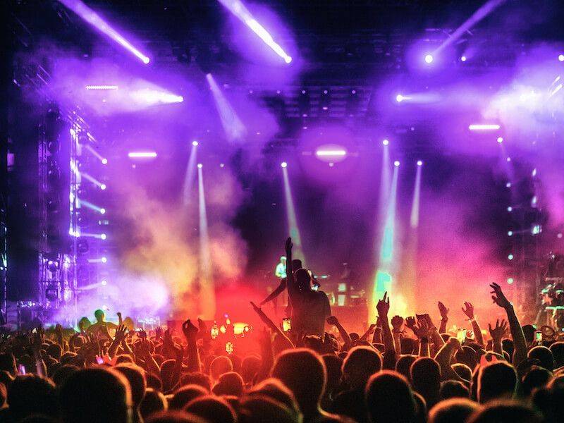 Crowd at a concert under purple lights and smoke, with raised hands and a performer silhouetted on stage