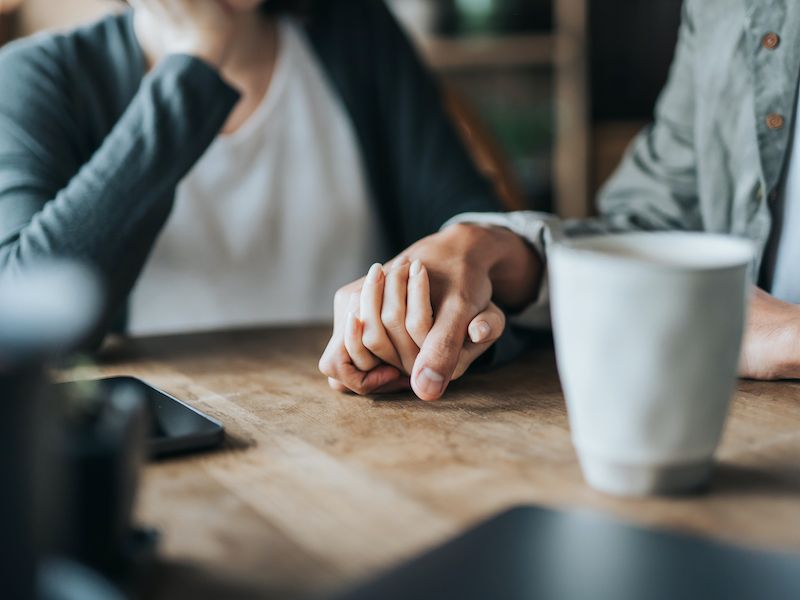 Two people holding hands across a wooden table beside a white coffee mug