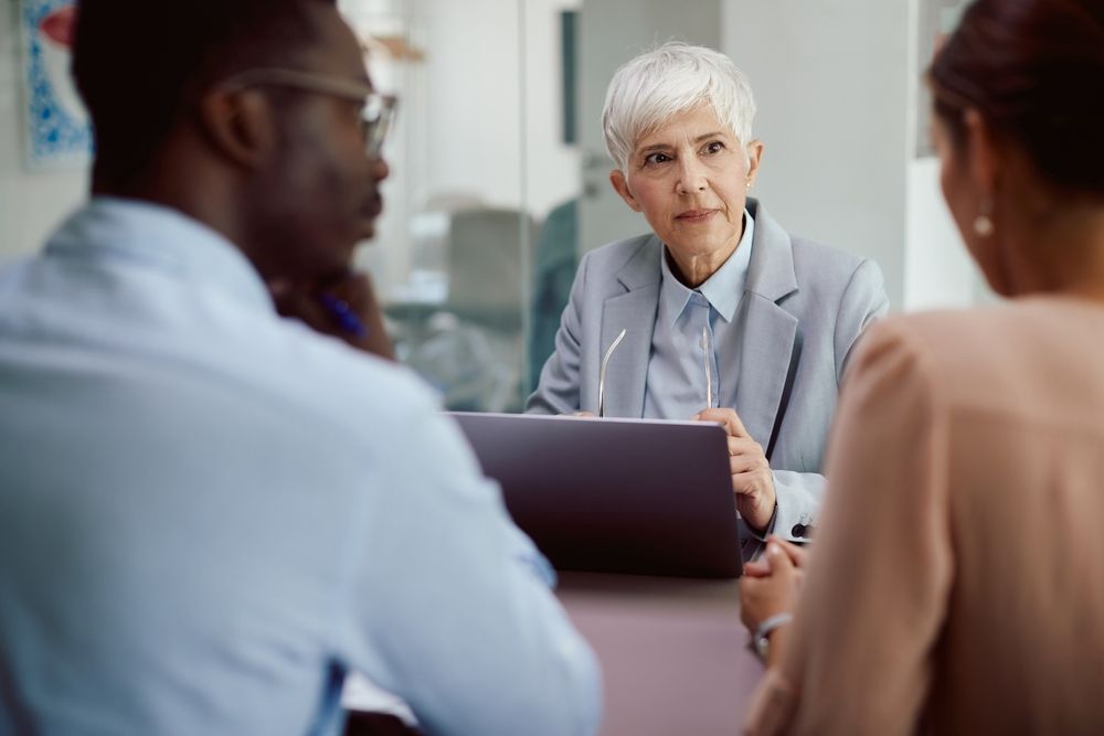 Three people in a meeting, with an older woman speaking behind a laptop.