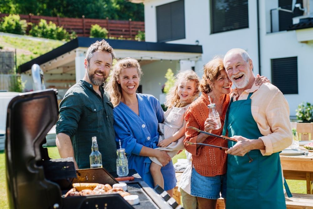 Group smiling by a barbecue grill in a sunny backyard, holding drinks and food.