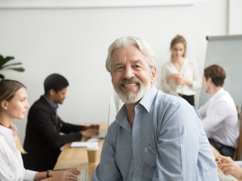 Smiling man in light blue shirt seated at a conference table with coworkers in a bright office