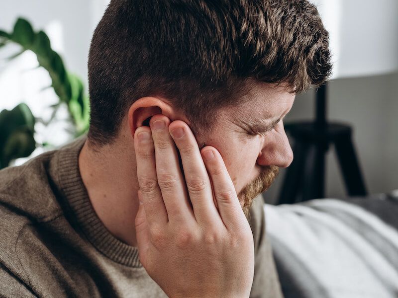 Man holding his ear and looking pained on a couch in a bright living room