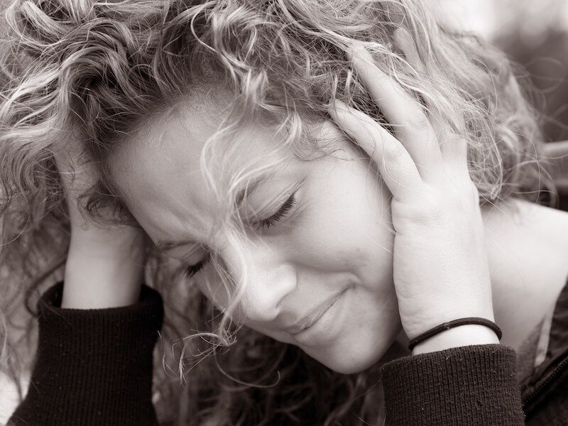 Close-up of a person with curly hair, eyes closed, holding their head with both hands in a pained expression