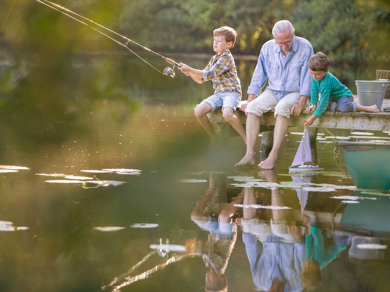 Three people sit on a dock, fishing with lines over a calm pond reflected in the water.