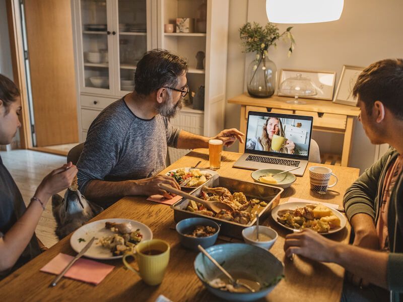 Friends share dinner over a video call at a dining table, with bowls of food and a laptop in a warm kitchen.