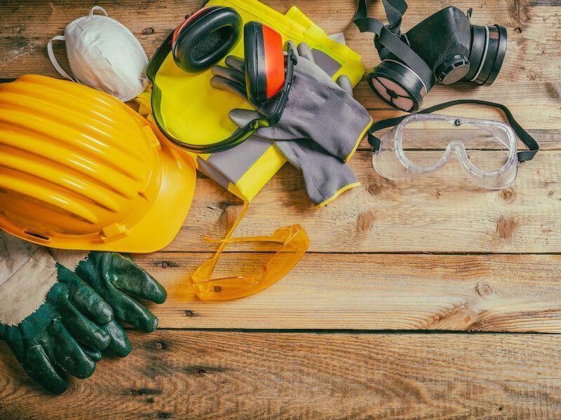 Safety gear on wooden table: yellow hard hat, gloves, goggles, earmuffs, dust mask, and knee pads