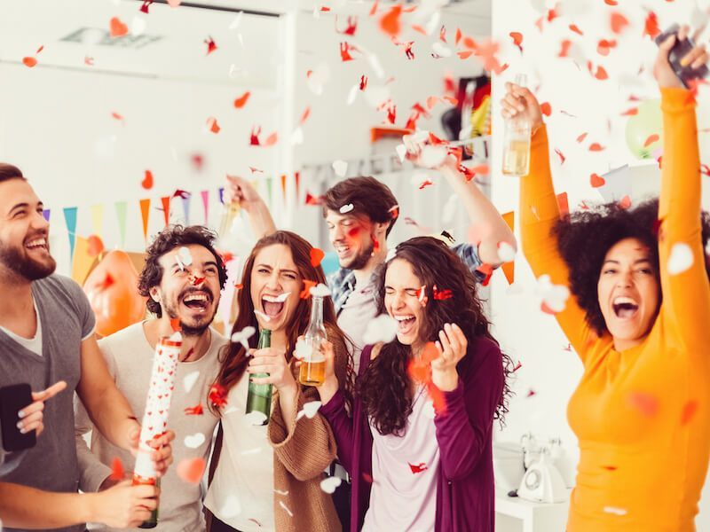 Friends celebrating indoors with confetti, smiling, cheering, and raising drinks at a party.