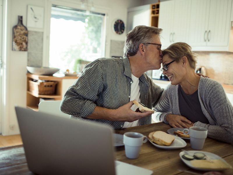 Two people sharing a kiss at a kitchen table with mugs, plates, and a laptop nearby