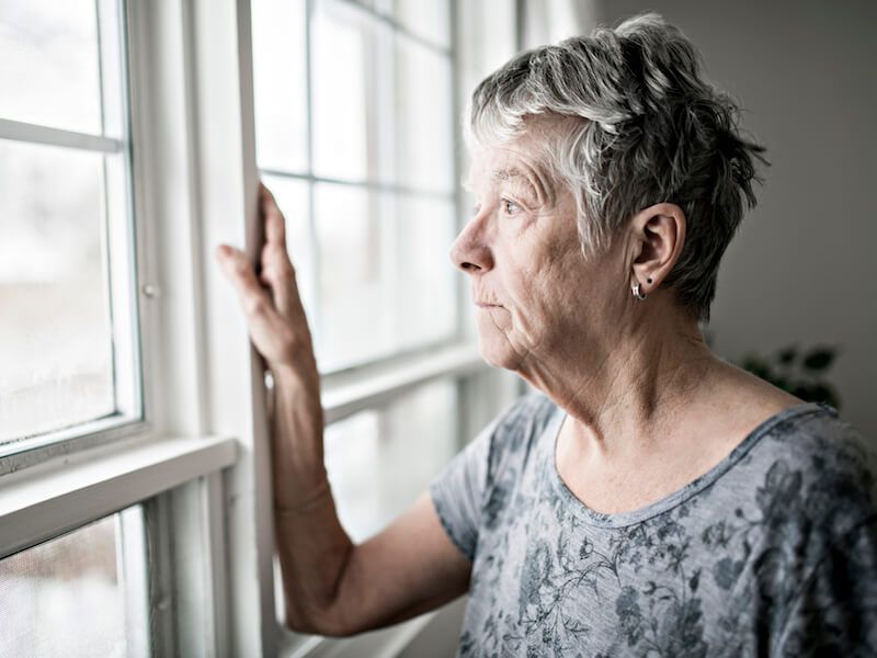 Elderly person gazing out a window, resting a hand on the frame in a bright room