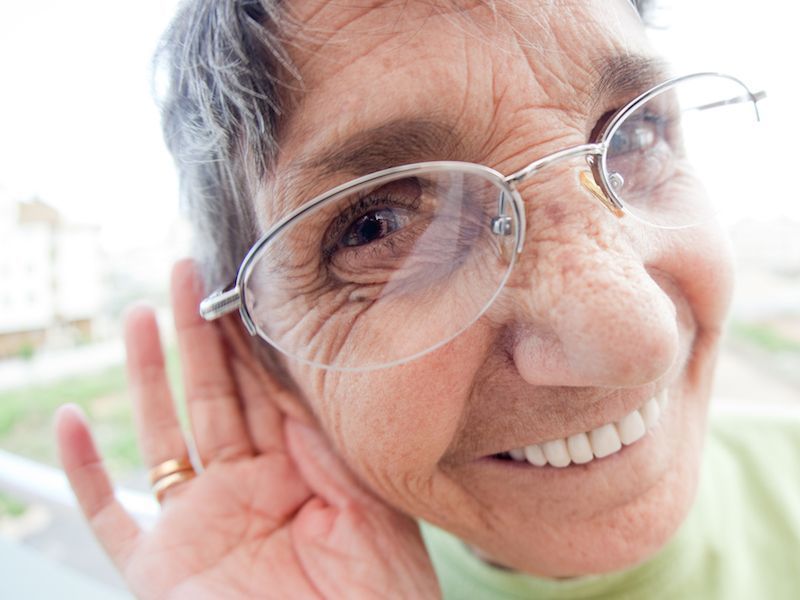 Close-up of a smiling person adjusting eyeglasses, hand cupped behind the ear, in bright light.