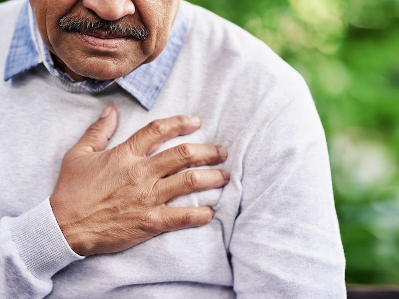 Man clutching his chest in a light gray sweater, suggesting chest pain or discomfort outdoors.