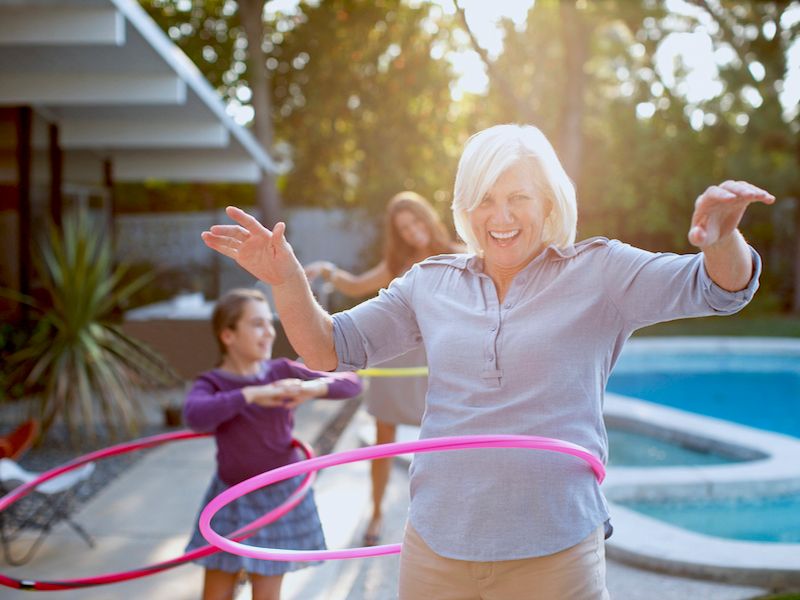 Woman with a pink hula hoop smiling in a backyard pool area, with a child in the background