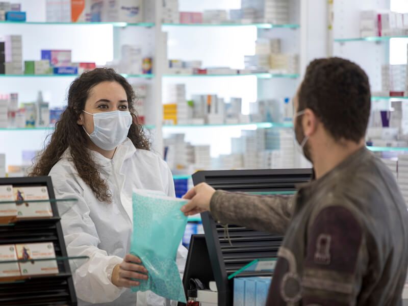 Masked pharmacist handing a turquoise bag to a customer at a pharmacy counter