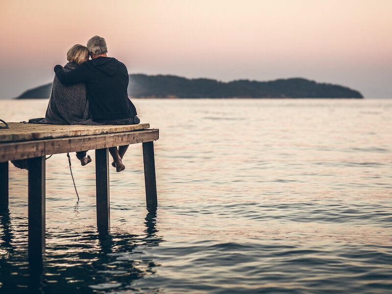 Two people sit on a wooden dock, hugging at sunset over calm water.