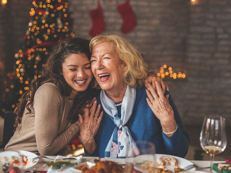 Two smiling women laugh together at a festive dinner table with candles and holiday lights in the background