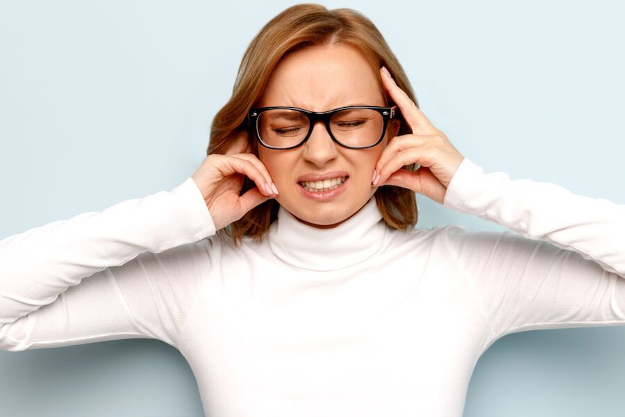 Woman in white turtleneck and glasses pressing her temples, looking frustrated on a light background