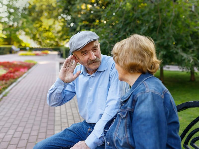 Two people sitting on a park bench, talking, with trees and flowers in the background.