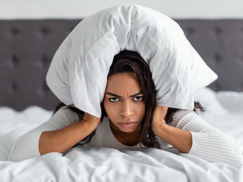 Person lying in bed with a pillow over their head, looking frustrated or tired.