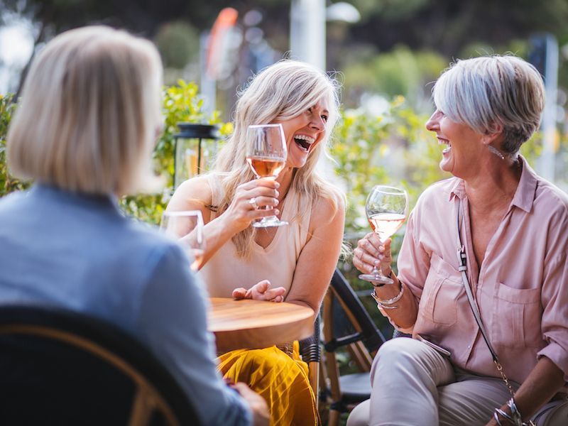 Three women chatting outdoors at a café, smiling and holding wine glasses.