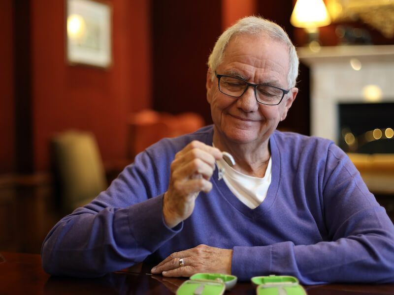 Person in a purple sweater smiling and holding a white face mask at a table in a warm indoor setting