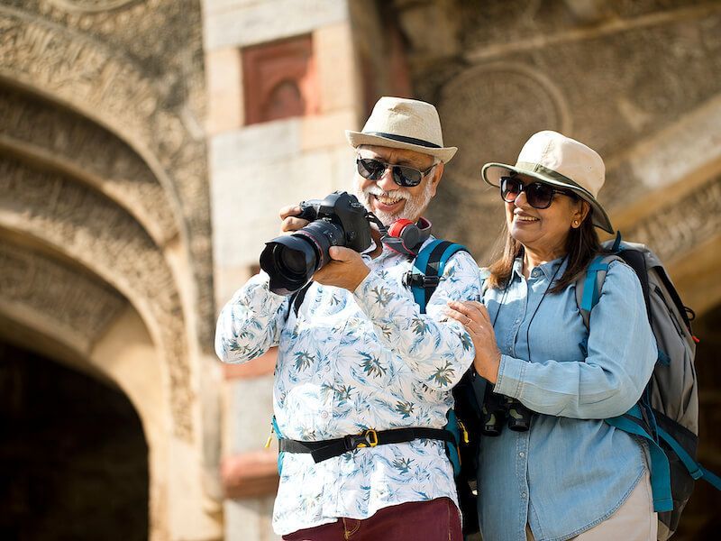 Two hikers with backpacks and cameras smiling in front of a stone bridge