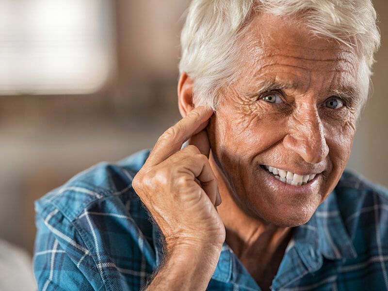 Smiling man in a blue plaid shirt touching his ear, seated indoors.