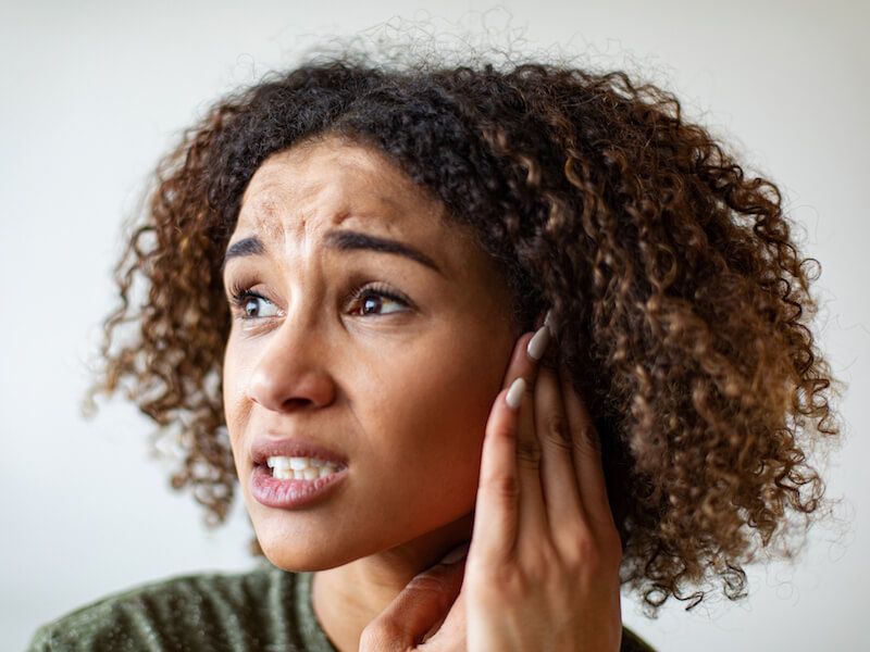 Woman holding her ear and wincing, looking worried against a light background