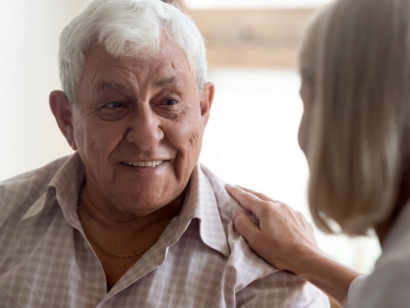 Older man smiling as a woman gently touches his shoulder indoors